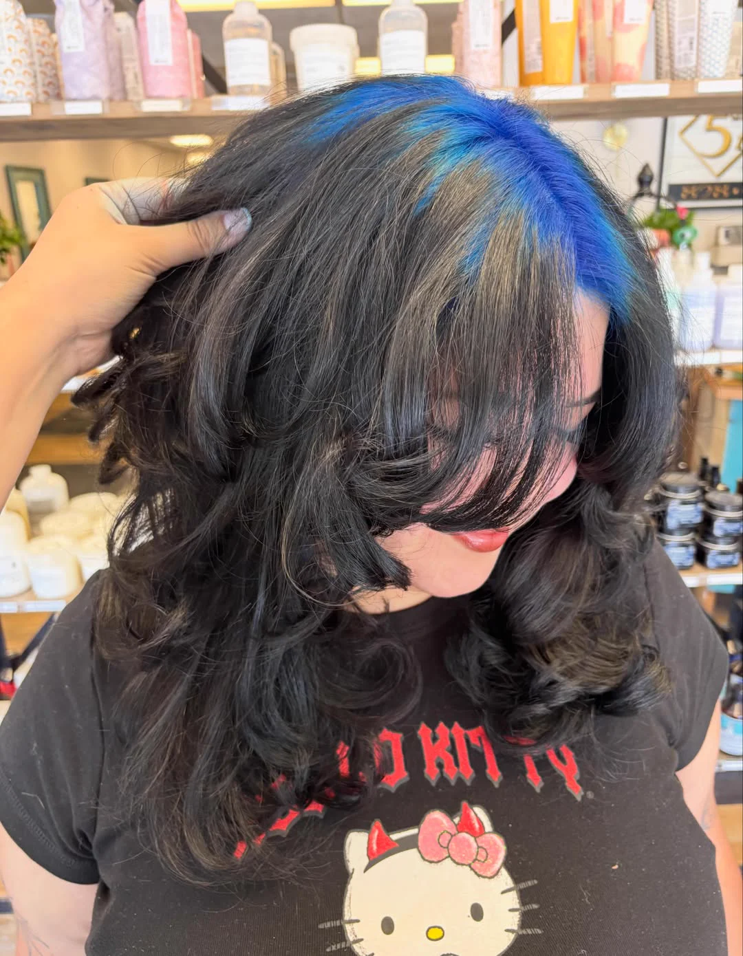 Woman with medium black wavy hair and electric-blue roots sweeping from a side part across the crown.