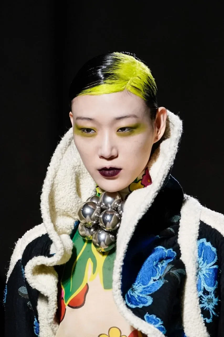 Woman with short slicked-back black hair and a broad neon-yellow wash across the front and crown roots.